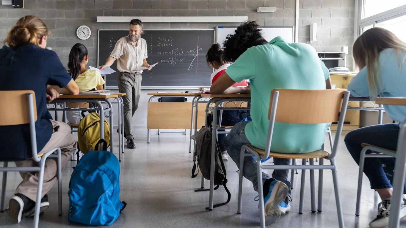 Mature caucasian man teacher hands out exams to multiracial high school students. Students ready to take exam.