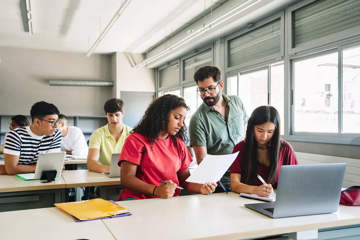 Friendly Professor explaining exercise to students in high school classroom. Young Teacher with beard helping teenager girl – Focus on Male teacher
